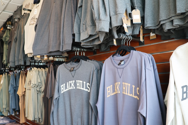 skygifts3 Clothing rack in a store showing gray and blue sweatshirts, front displays say 'BLACK HILLS'.